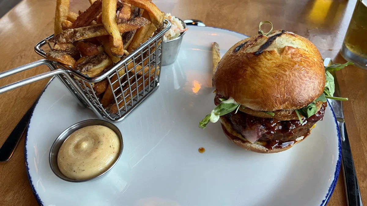 A burger in a toasted bun with greens, a metal basket of crispy fries, and a small cup of aioli on a white plate
