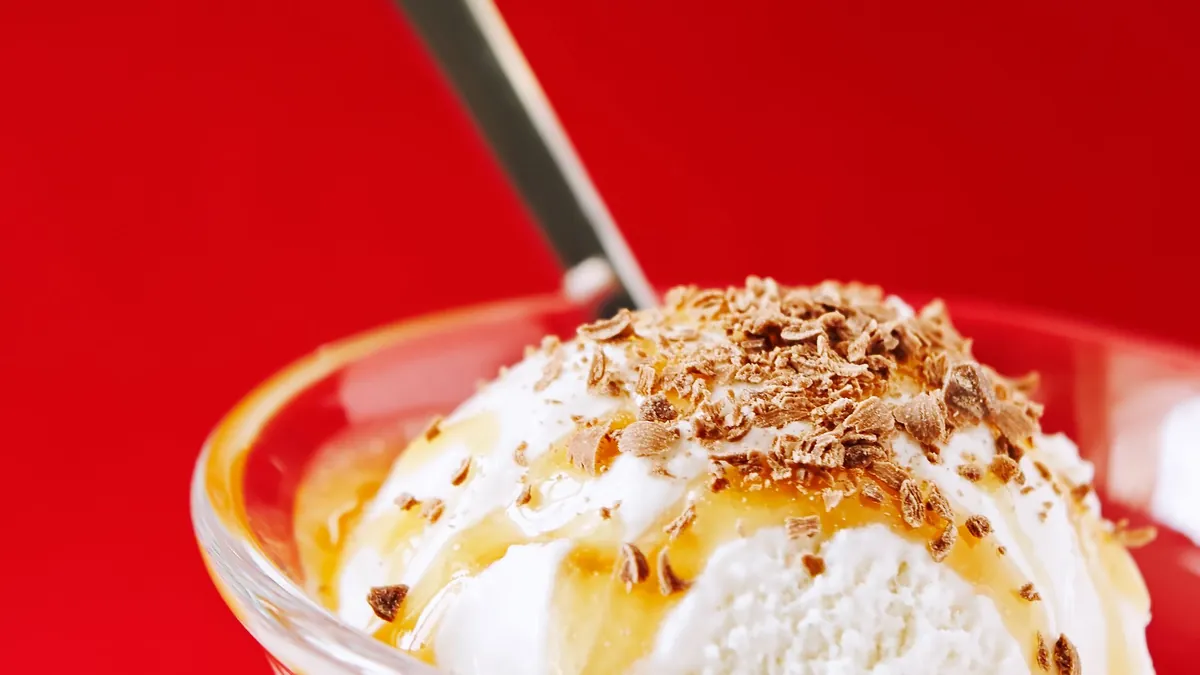 A scoop of vanilla ice cream with caramel drizzle and chocolate shavings served in a glass dish, with a spoon.