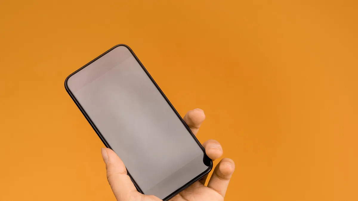 A hand holding a smartphone with a blank screen against an orange background.