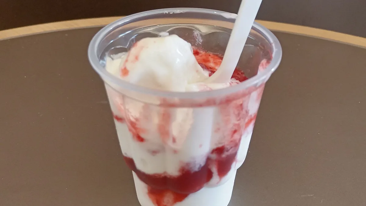A clear plastic cup of soft serve swirled with strawberry topping and a white spoon